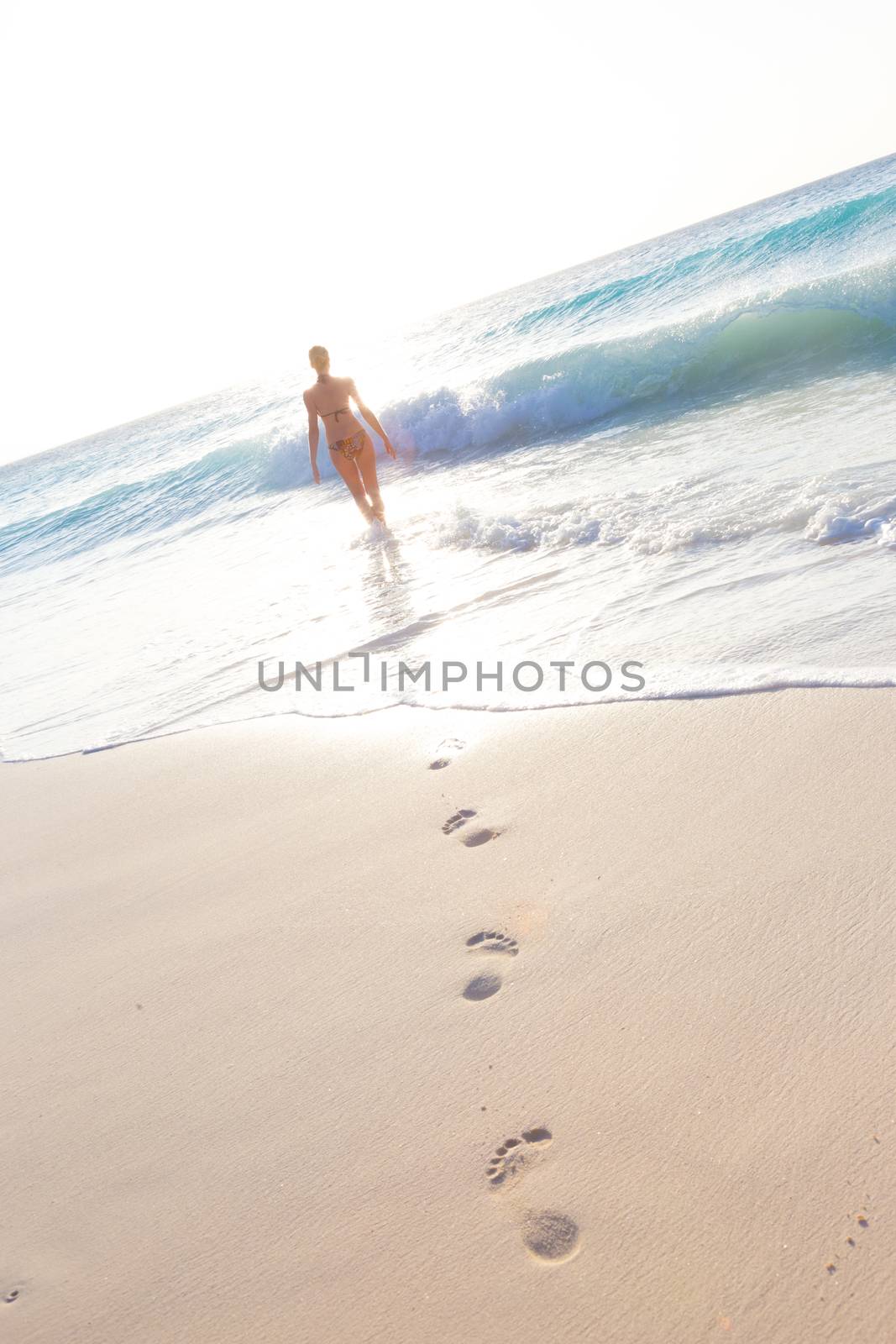 Happy woman enjoying in summer, running joyfully on tropical beach in sunset. Beautiful caucasian model wearing bikini on vacations on sandy beach. Footprints in sand. 