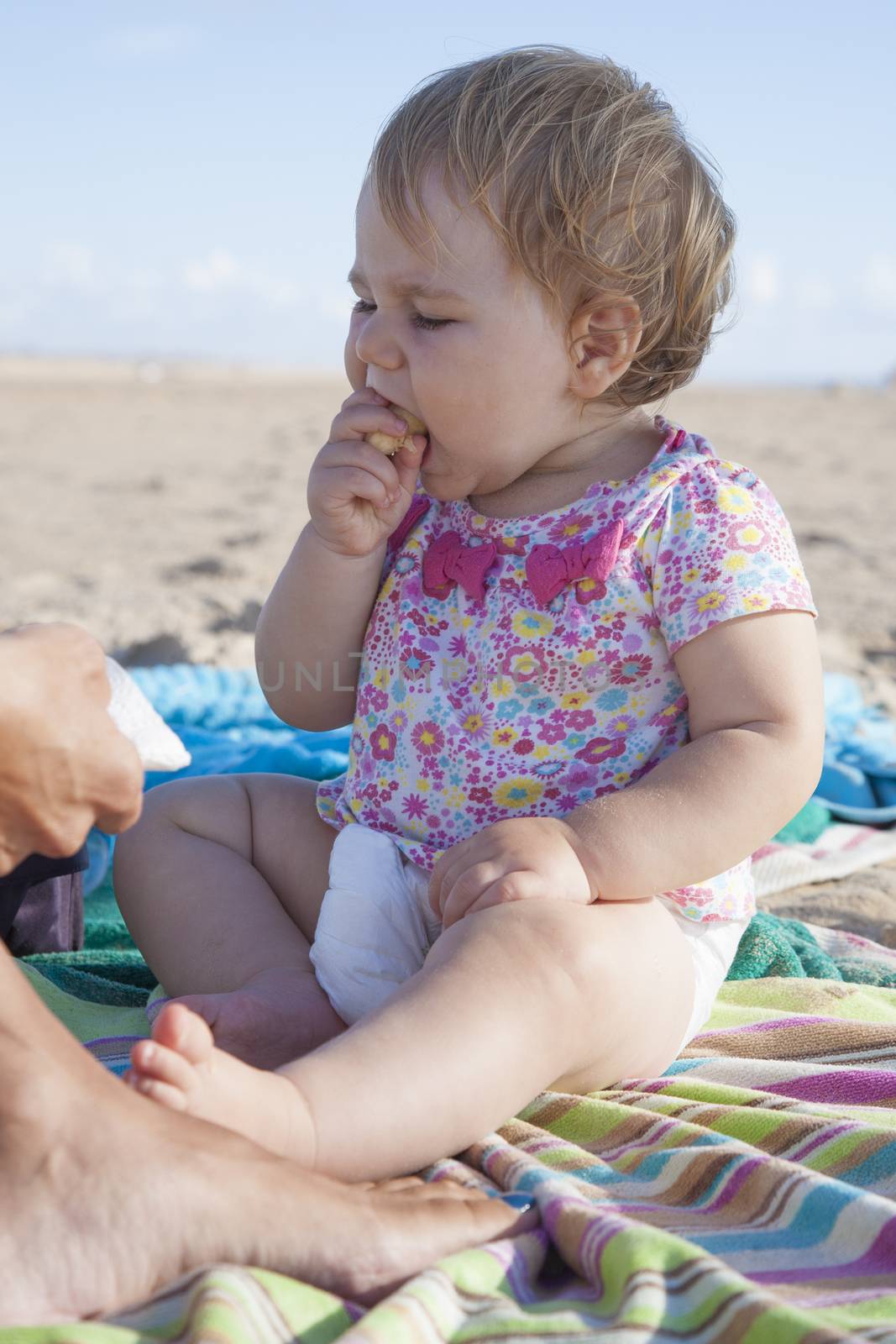 portrait of one year baby eating banana on towel next to mom at beach