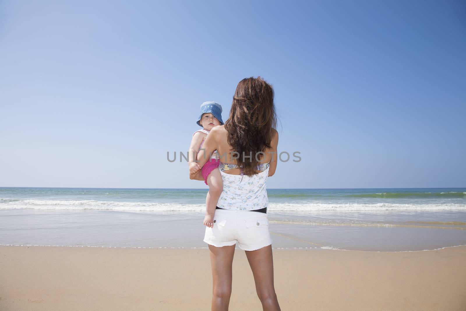 one year blonde baby in brunette woman mother arms back at beach Conil Cadiz Spain