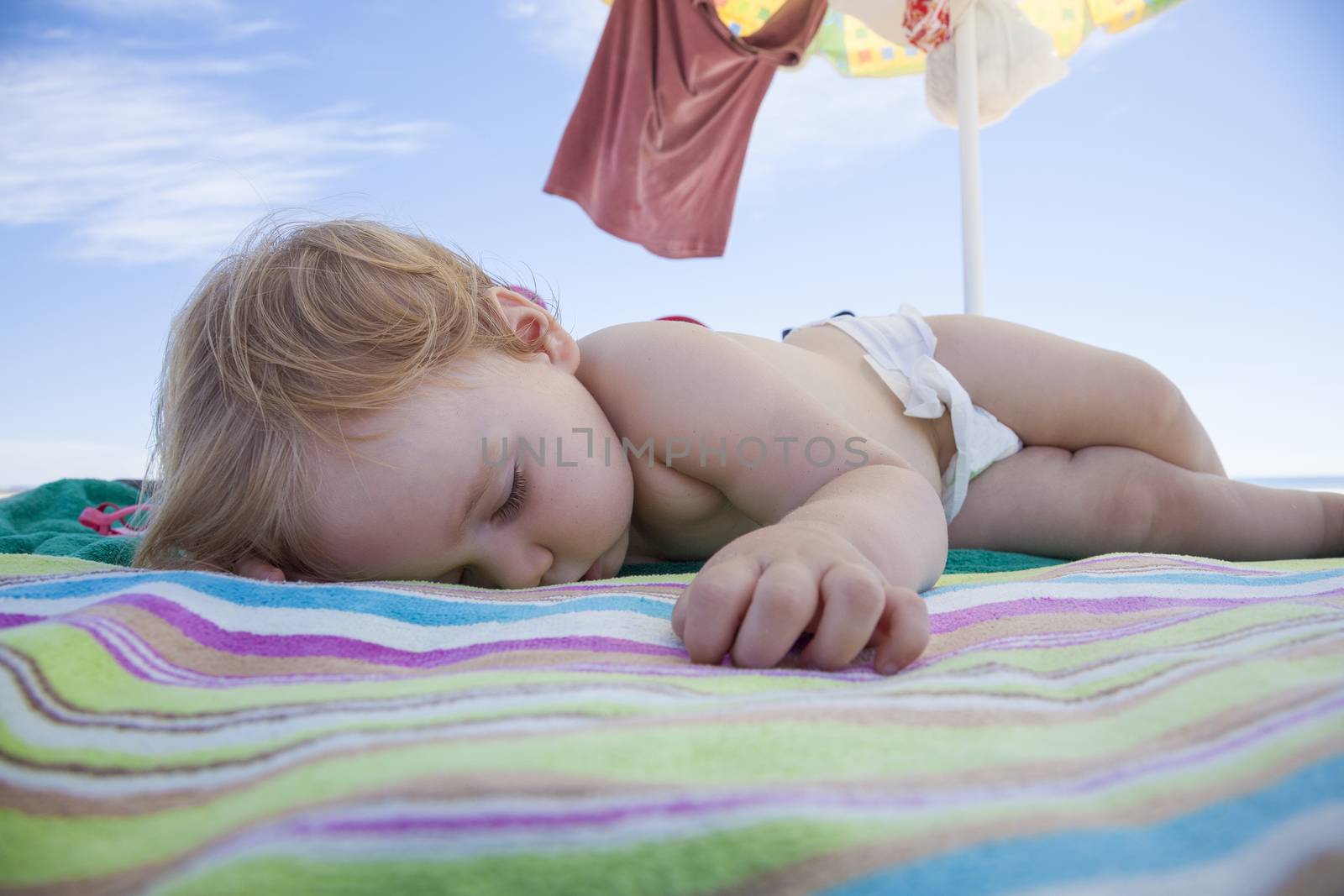 one year baby with white diaper sleeping on striped colored beach towel under parasol