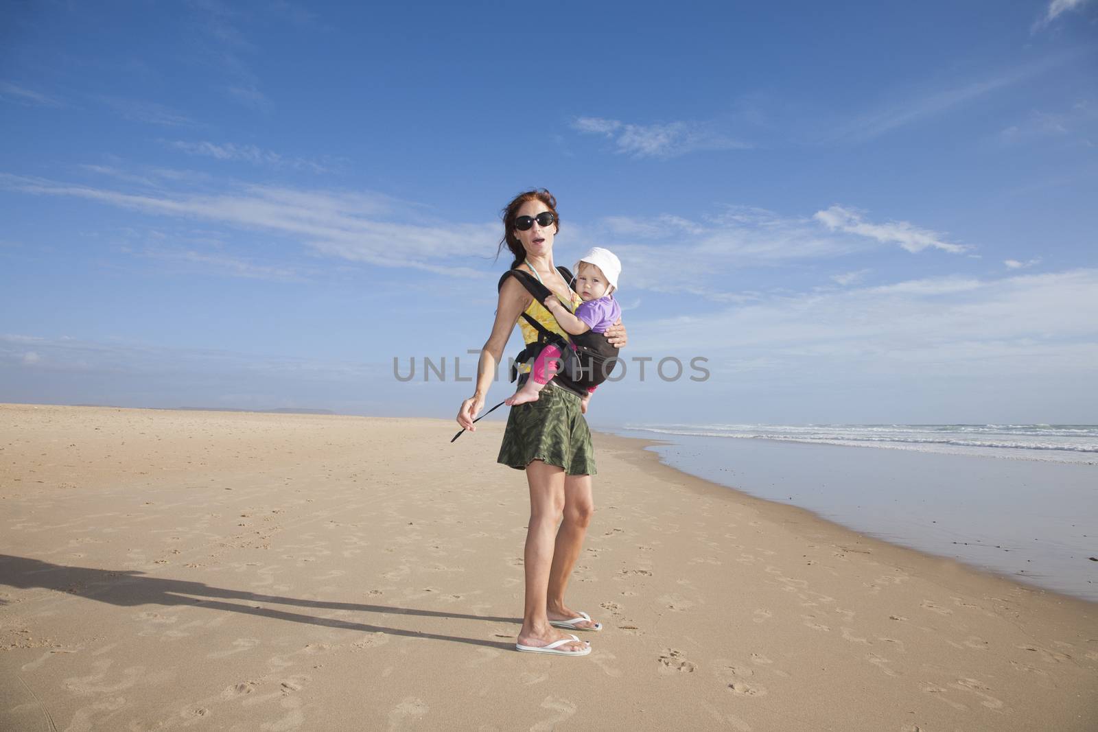 mother with green miniskirt black sunglasses carrying her one year baby with white hat in front rucksack walking at a beach
