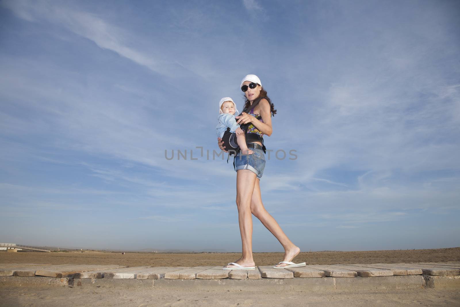 mother with white cap black sunglasses blue jeans shorts carrying her one year baby with hat in front rucksack walking at a beach