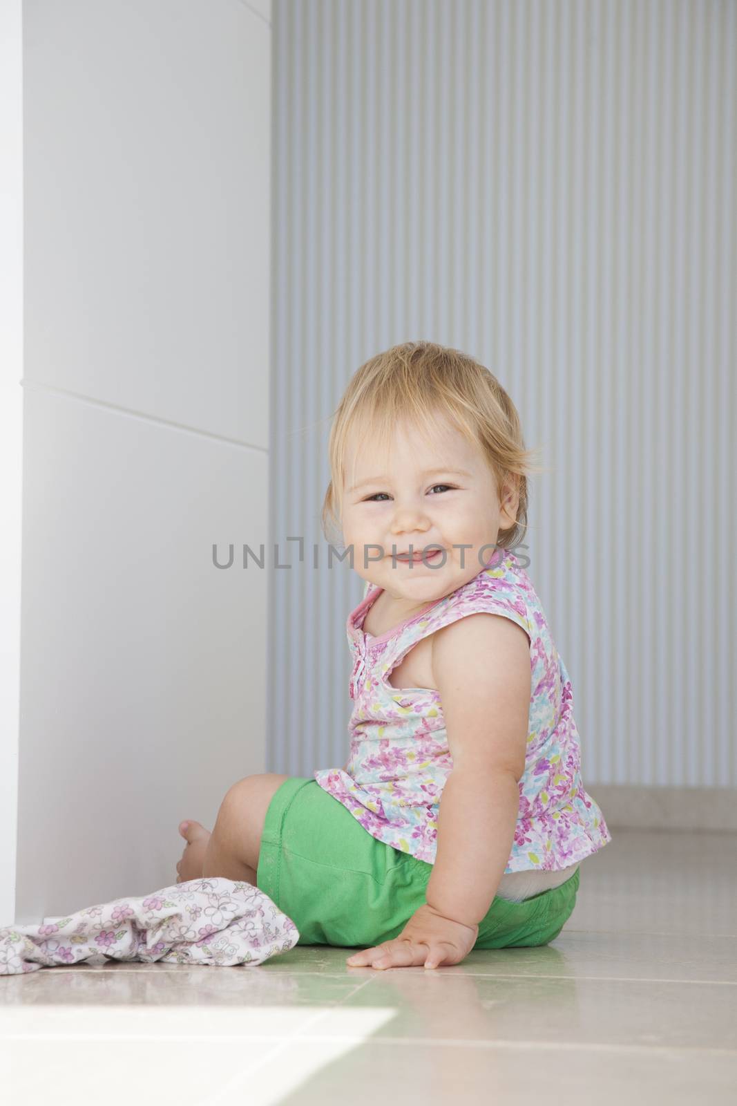 happy blonde one year baby green shorts smiling sitting on grey white floor