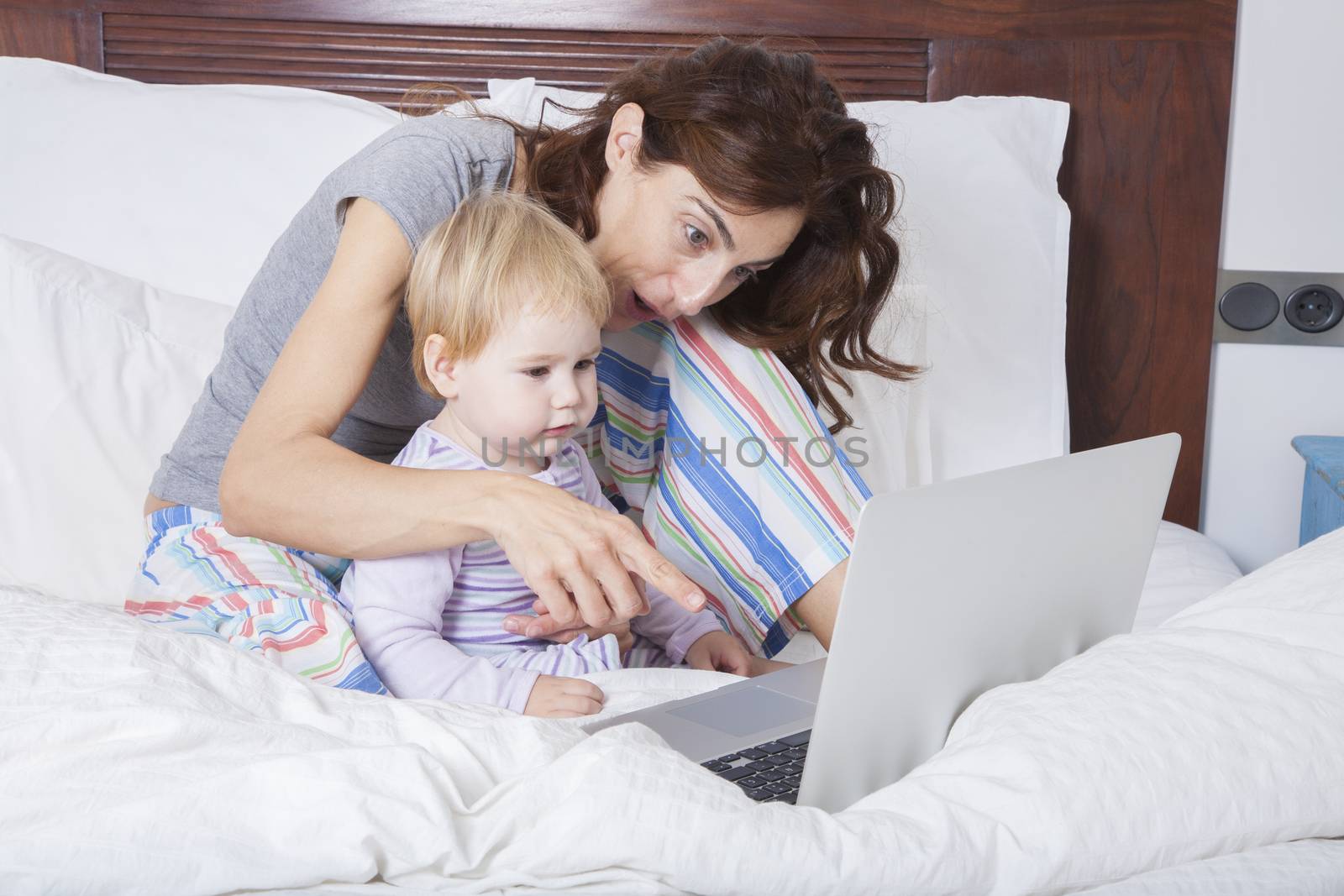 eighteen month aged blonde baby with brunette woman mother reading grey laptop on white bed