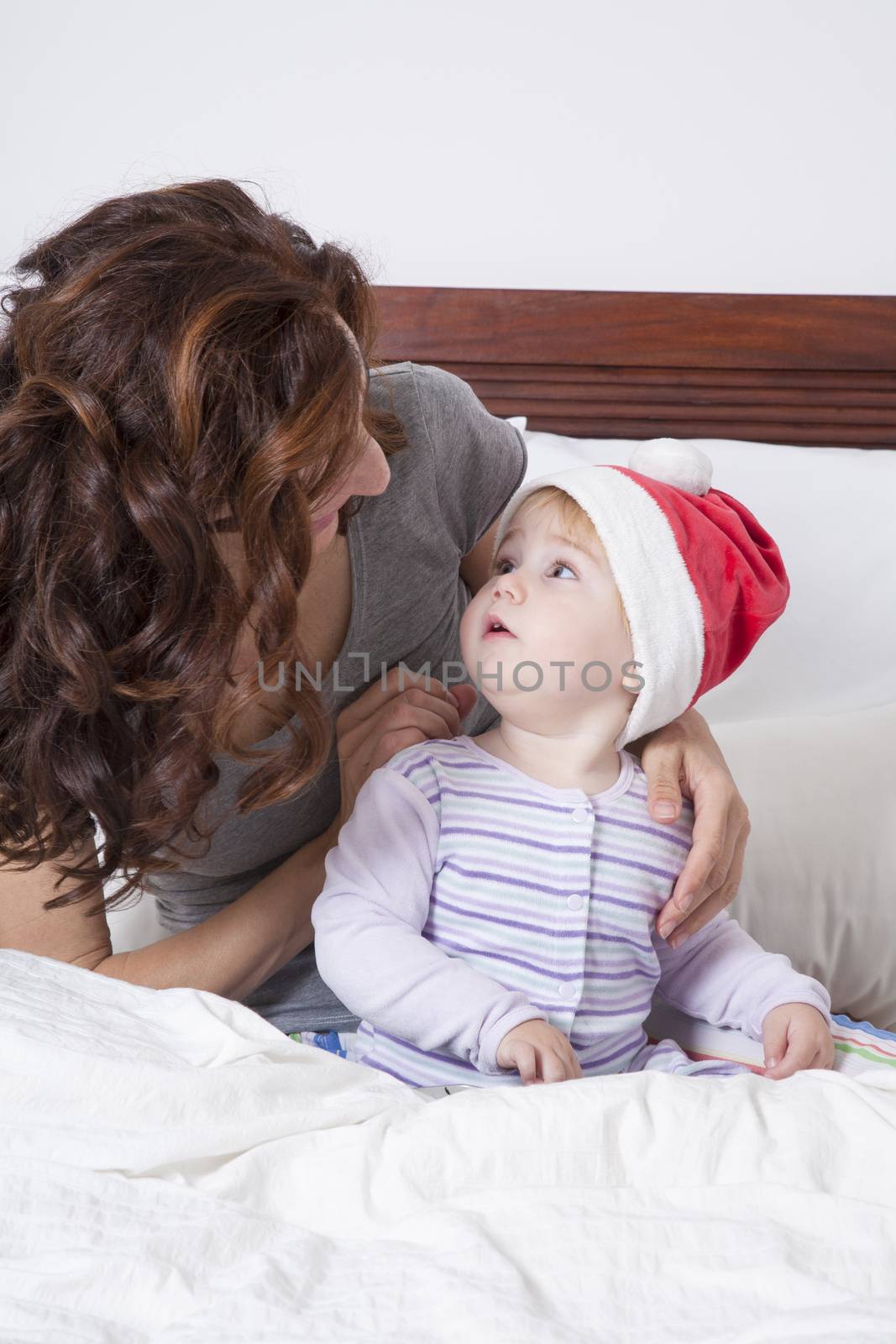 eighteen month aged blonde baby with Santa Claus hat and brunette woman mother on white bed