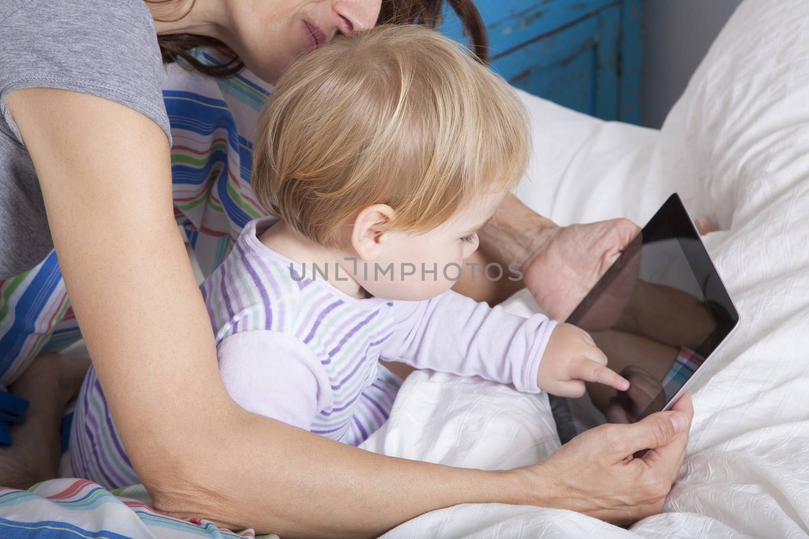 eighteen month aged blonde baby with brunette woman mother reading digital tablet on white bed