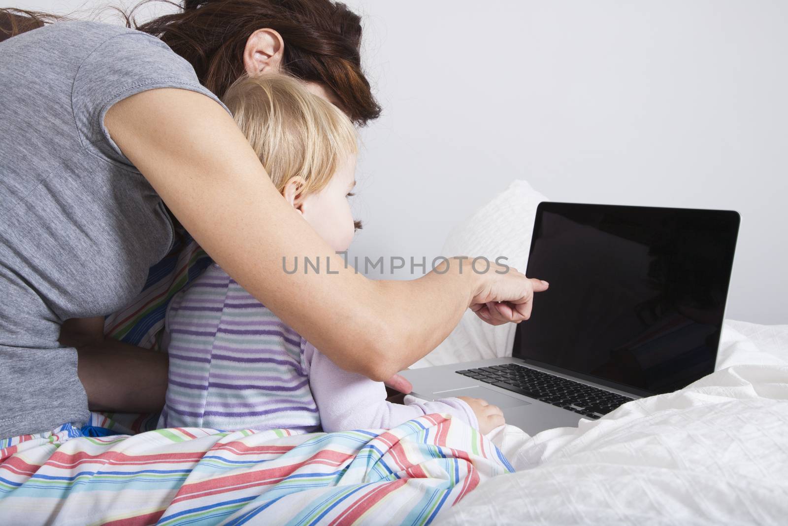 eighteen month aged blonde baby with brunette woman mother reading grey laptop on white bed