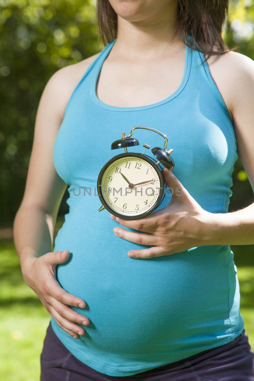 young pregnant woman blue shirt with clock in her hands over green tree background
