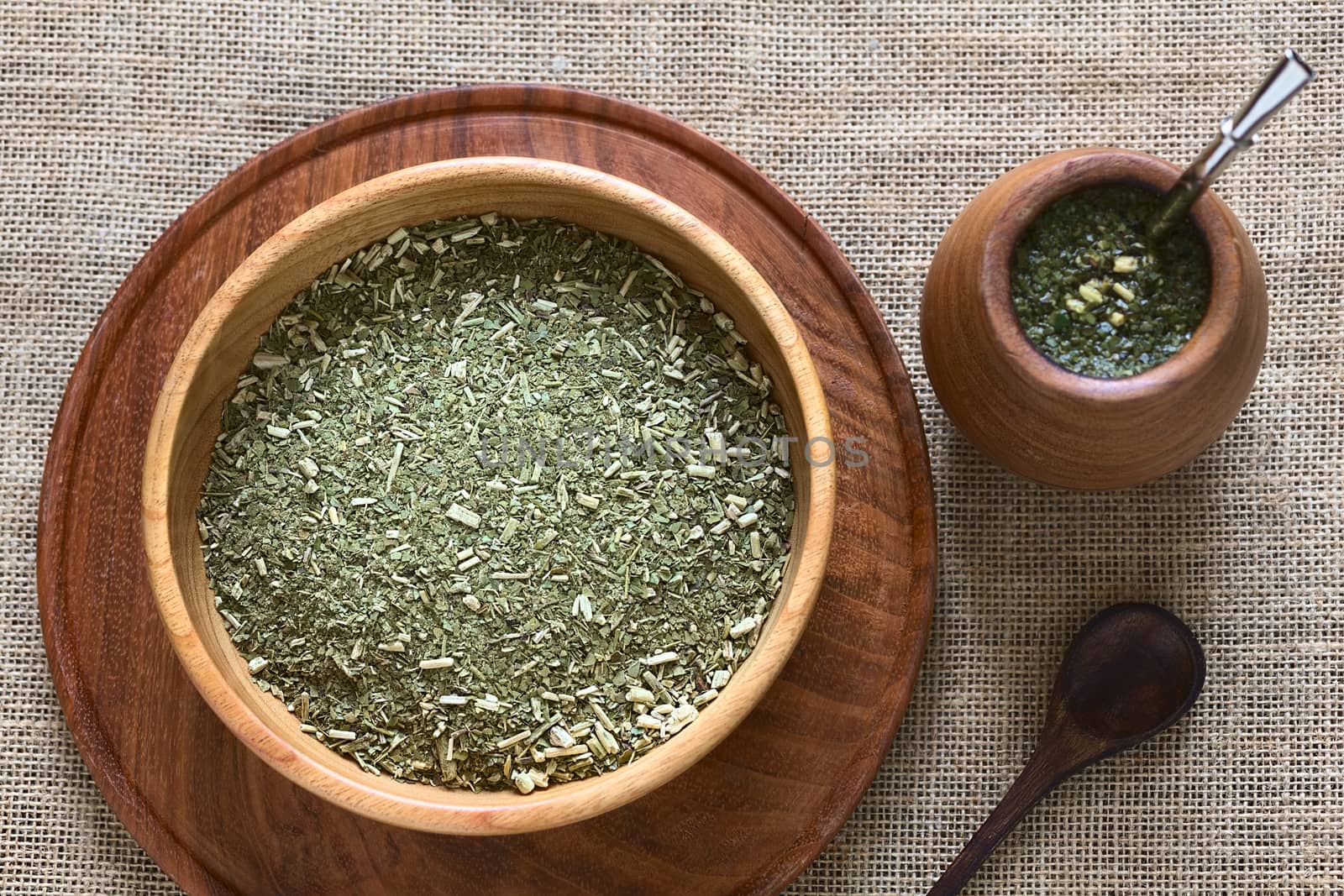 South American yerba mate (mate tea) dried leaves in wooden bowl with a wooden mate cup and bombilla (straw) filled with tea next to it photographed with natural light. Mate is the national infusion of Argentina. (Selective Focus, Focus on the dried tea leaves) 