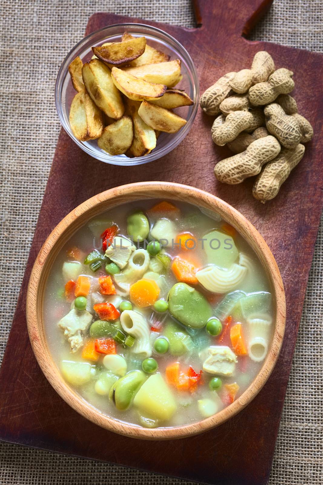 Overhead shot of Bolivian traditional Sopa de Mani (peanut soup) made of meat, pasta, vegetables (pea, carrot, potato, broad bean, pepper, corn) with ground peanut in wooden bowl, traditionally served with fried potatoes, photographed with natural light (Selective Focus, Focus on the soup)