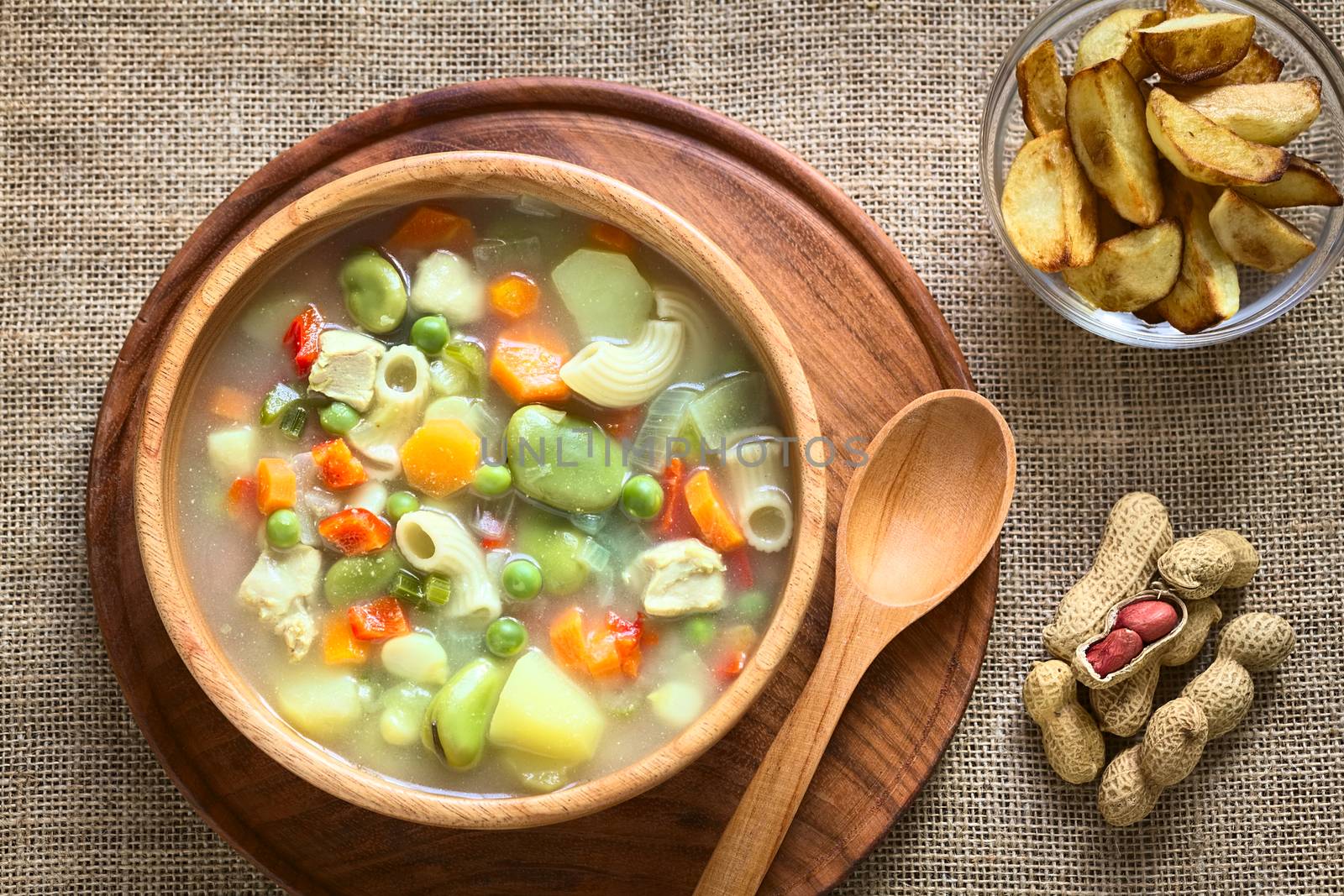 Overhead shot of Bolivian traditional Sopa de Mani (peanut soup) made of meat, pasta, vegetables (pea, carrot, potato, broad bean, pepper, corn) with ground peanut in wooden bowl, traditionally served with fried potatoes, photographed with natural light 