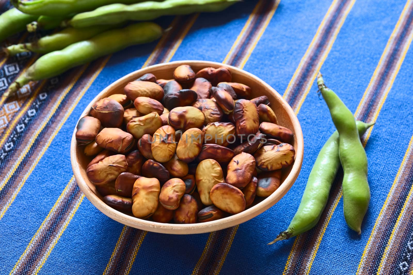 Roasted broad beans (lat. Vicia faba) eaten as snack in Bolivia in bowl with fresh broad bean pods on the side and in the back on blue fabric, photographed with natural light (Selective Focus, Focus in the middle of the roasted beans)