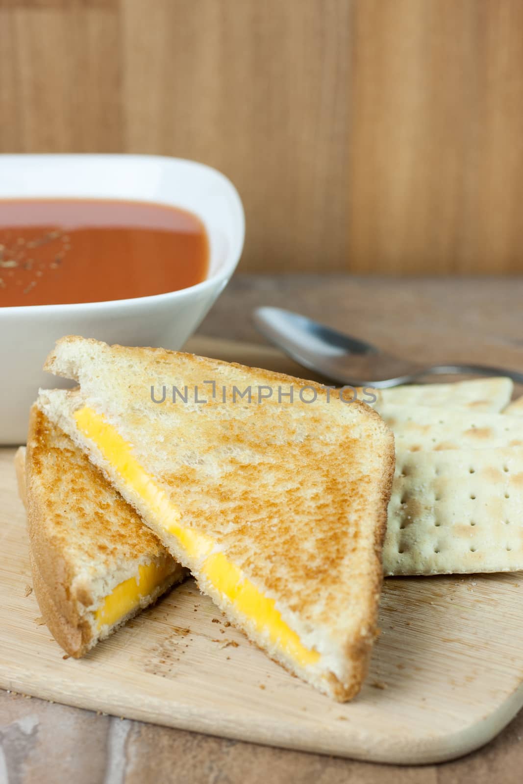 A grilled cheese sandwich with a bowl of tomato soup and saltine crackers on a wooden serving board.