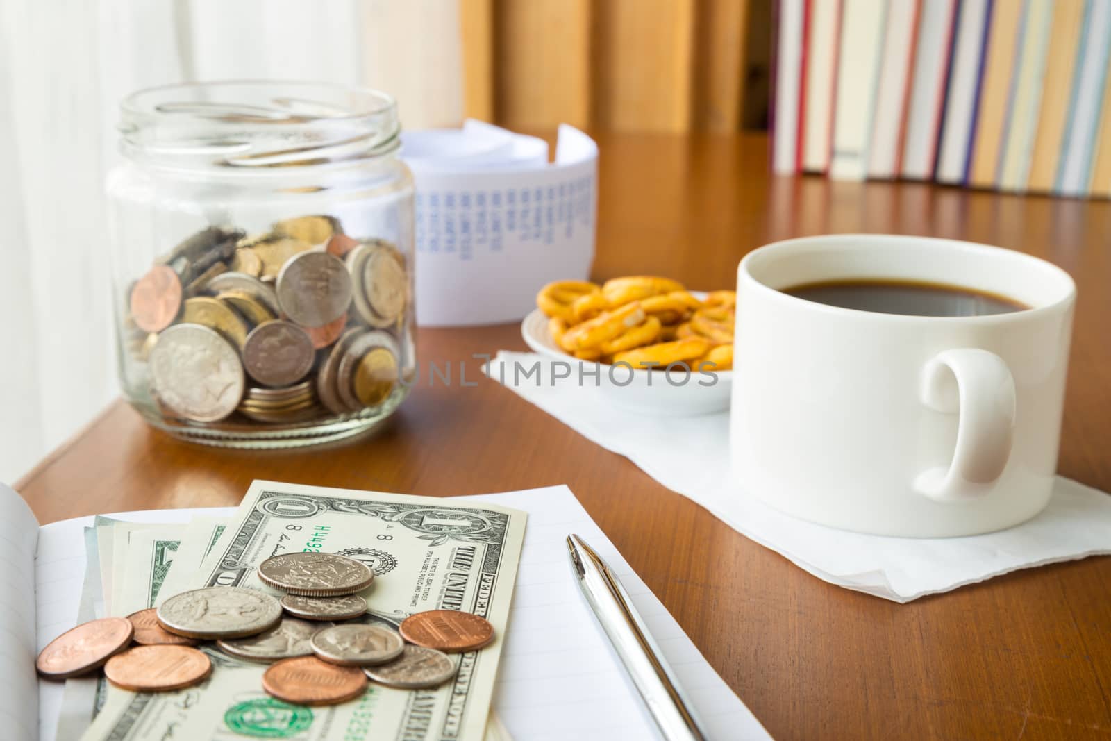 Many coins and banknotes place on notebook with a money saving jar, bills and coffee break on table, home finance concept