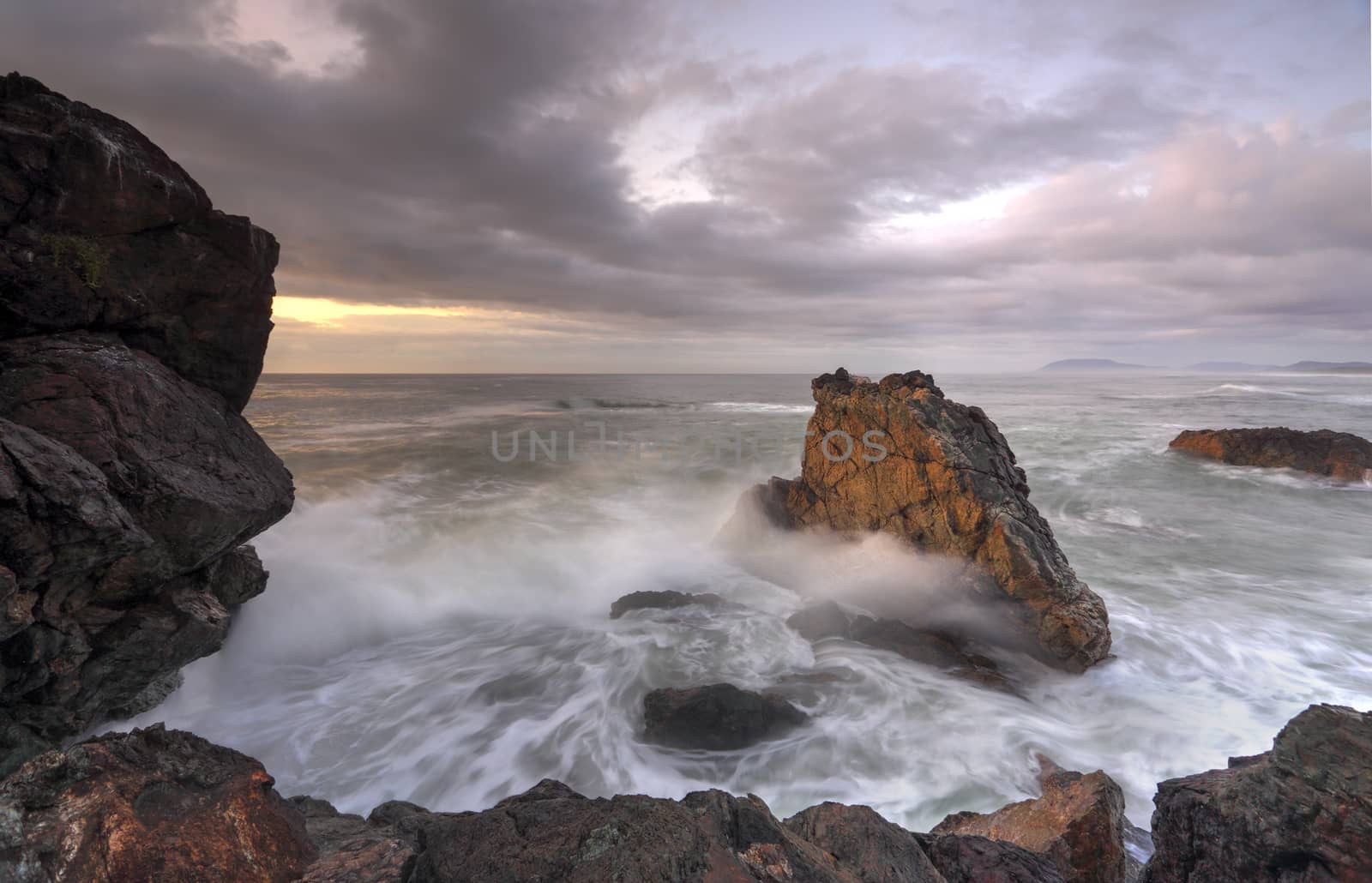 Sunrise at Lighthouse beach a soft golden glow warms the side of the rocks and highlights the water and clouds in a subtle way