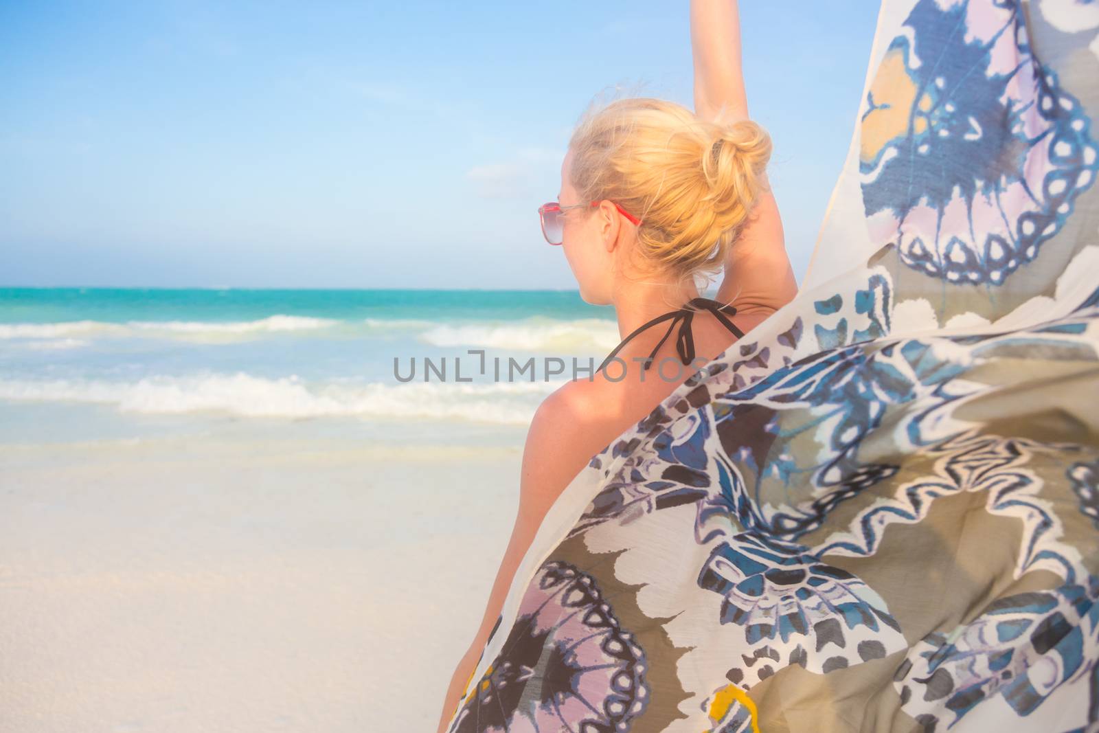 Happy woman enjoying, relaxing joyfully in summer by tropical blue water. Her colorful kerchief fluttering in wind on picture perfect Paje beach, Zanzibar, Tanzania.