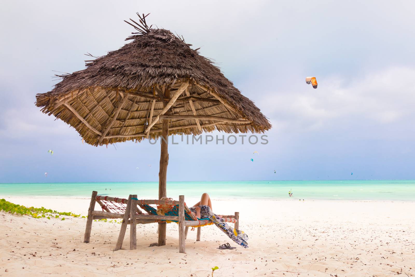 Women sunbathing on dack chair under wooden umbrella on stunning tropical beach. Kiteboarders turquoise blue lagoon of Paje beach, Zanzibar, Tanzania in the background.