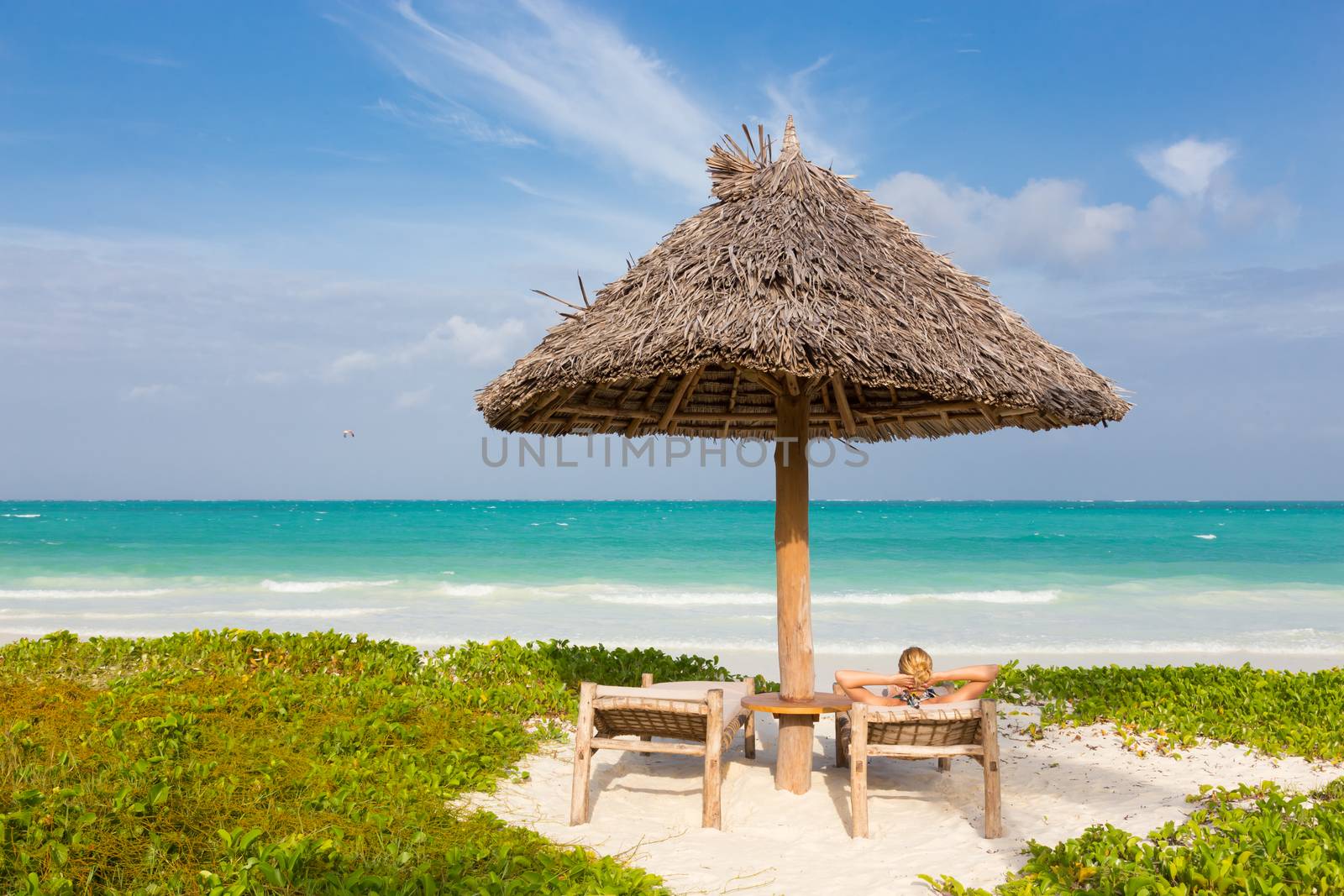 Women sunbathing on dack chair under wooden umbrella on stunning tropical beach. Turquoise blue lagoon of Paje beach, Zanzibar, Tanzania in the background.