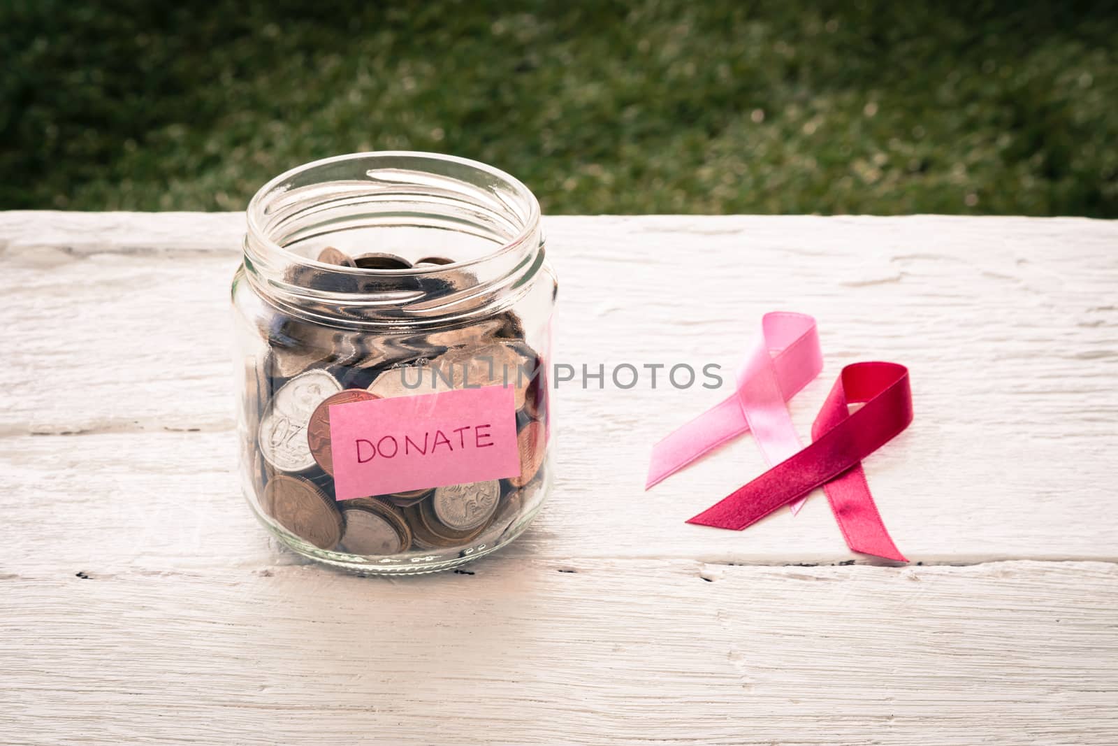 World coins in money glass jar with pink DONATE word label place on white wood table with two breast cancer ribbon signs, vintage and retro style image with vignette