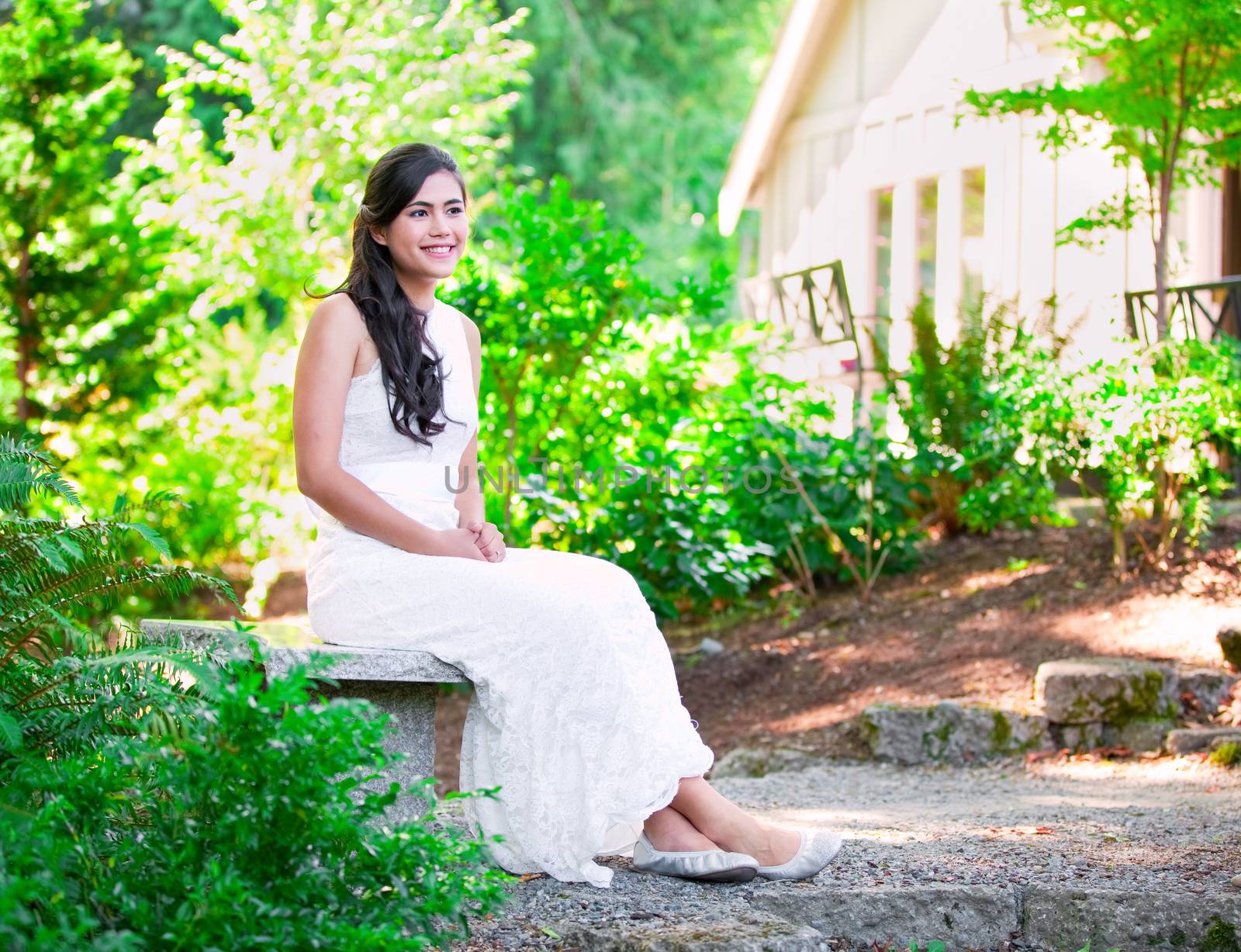 Beautiful biracial bride in white lace wedding dress sitting on bench outdoors, smiling