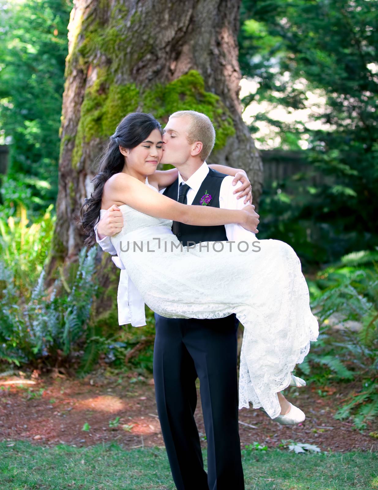 Caucasian groom carrying his biracial bride outdoors, with a kiss