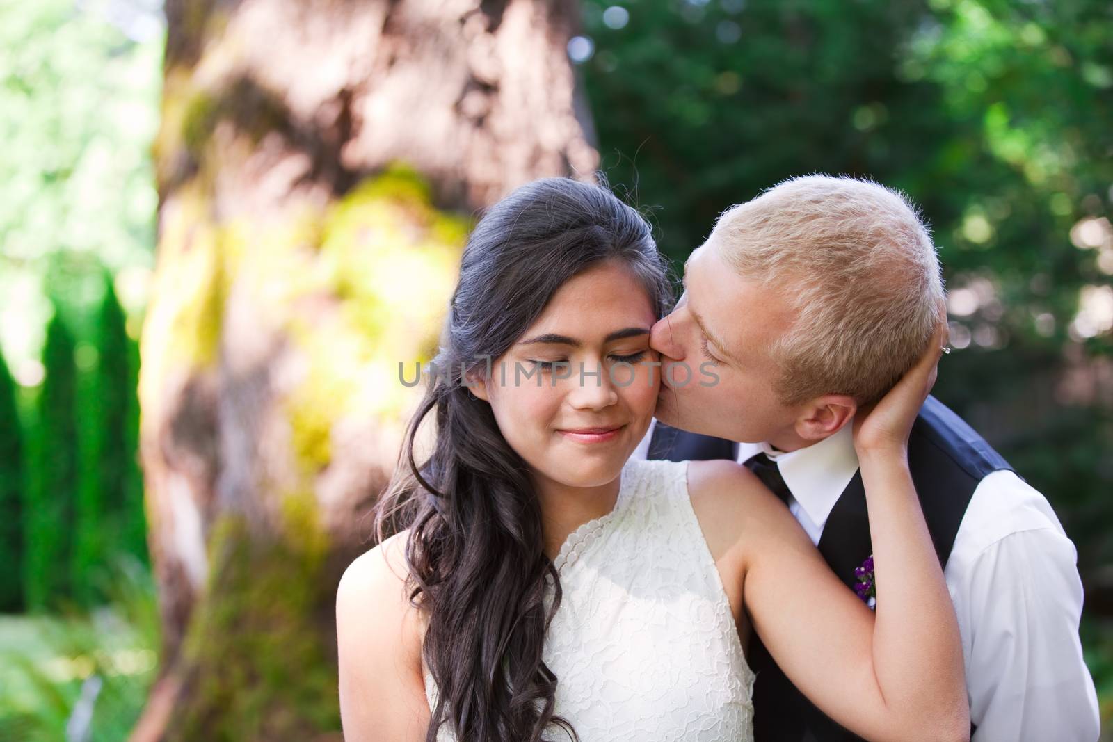 Caucasian groom lovingly kissing his biracial bride on cheek. Diverse couple
