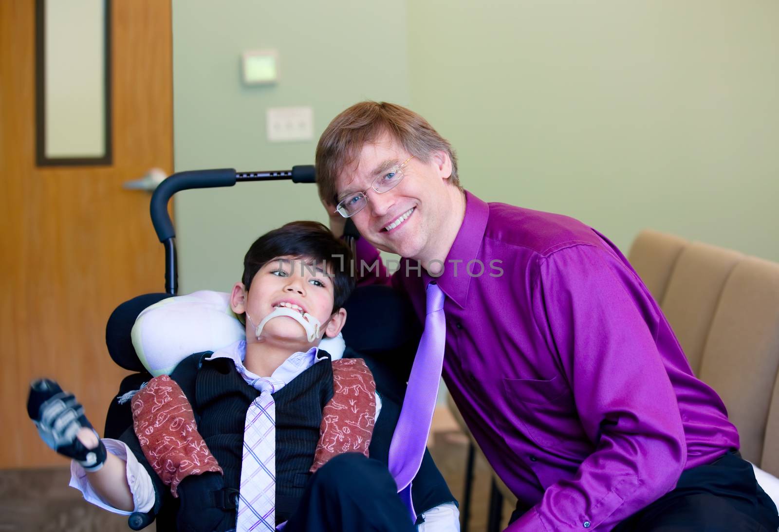 Caucasian father in purple dresshirt and necktie sitting next to biracial disabled son in wheelchair