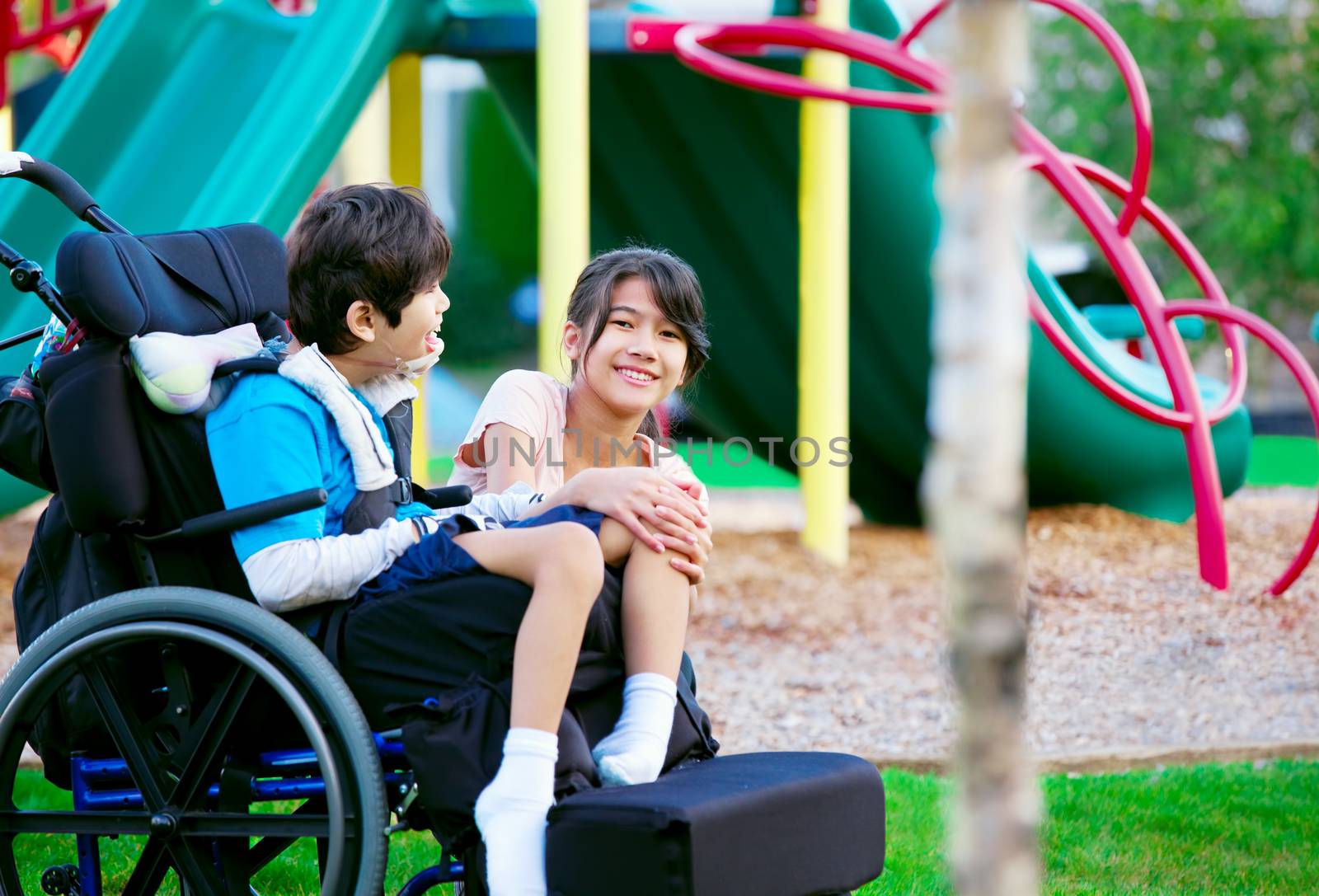 Sister sitting next to disabled brother in wheelchair at playground