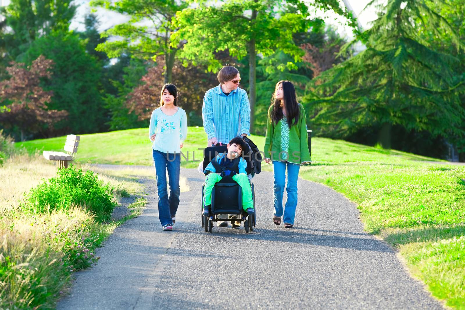 Disabled boy in wheelchair walking with family outdoors on sunny day in park