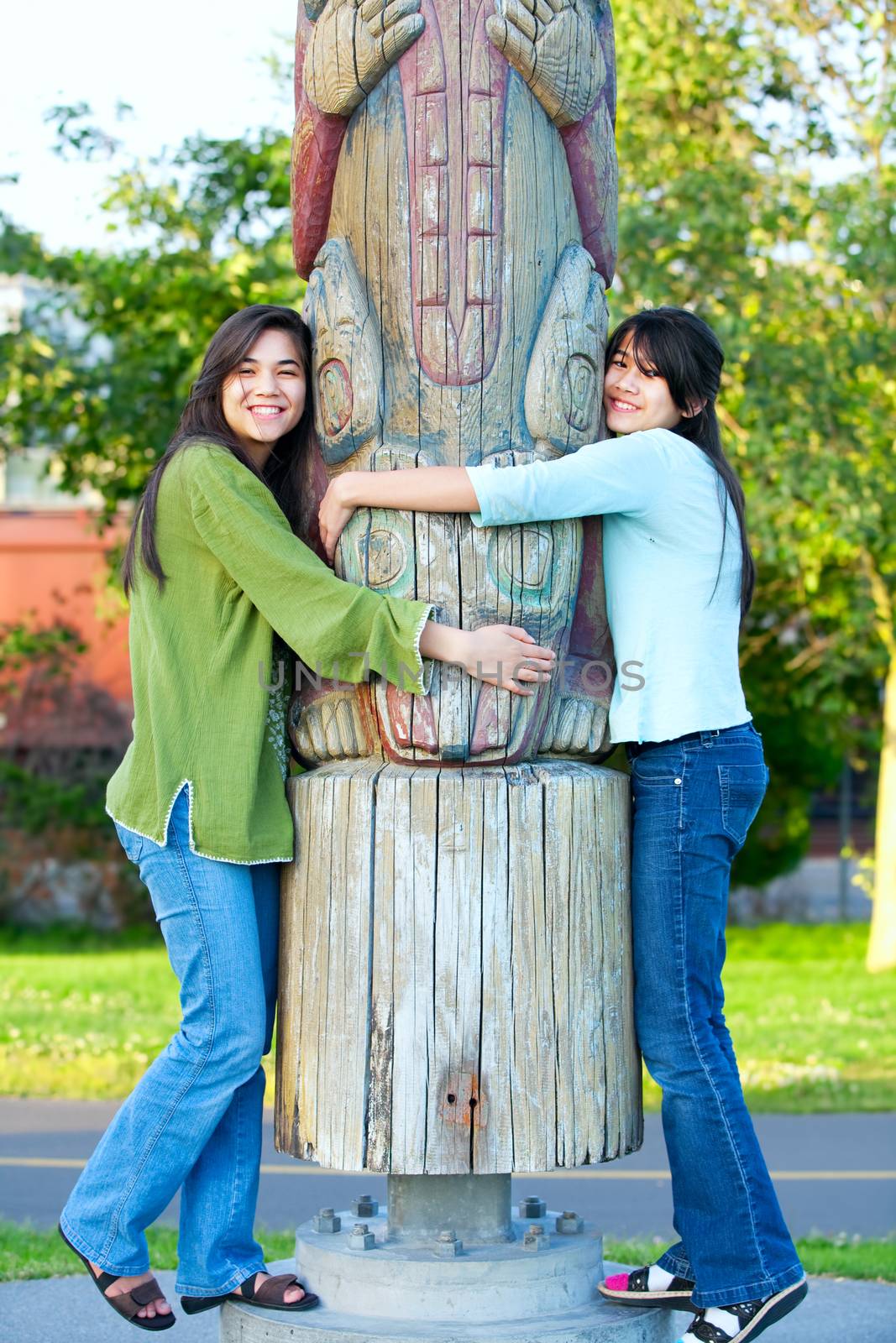 Two young, biracial teen girl in park hugging a totem pole on su by jarenwicklund