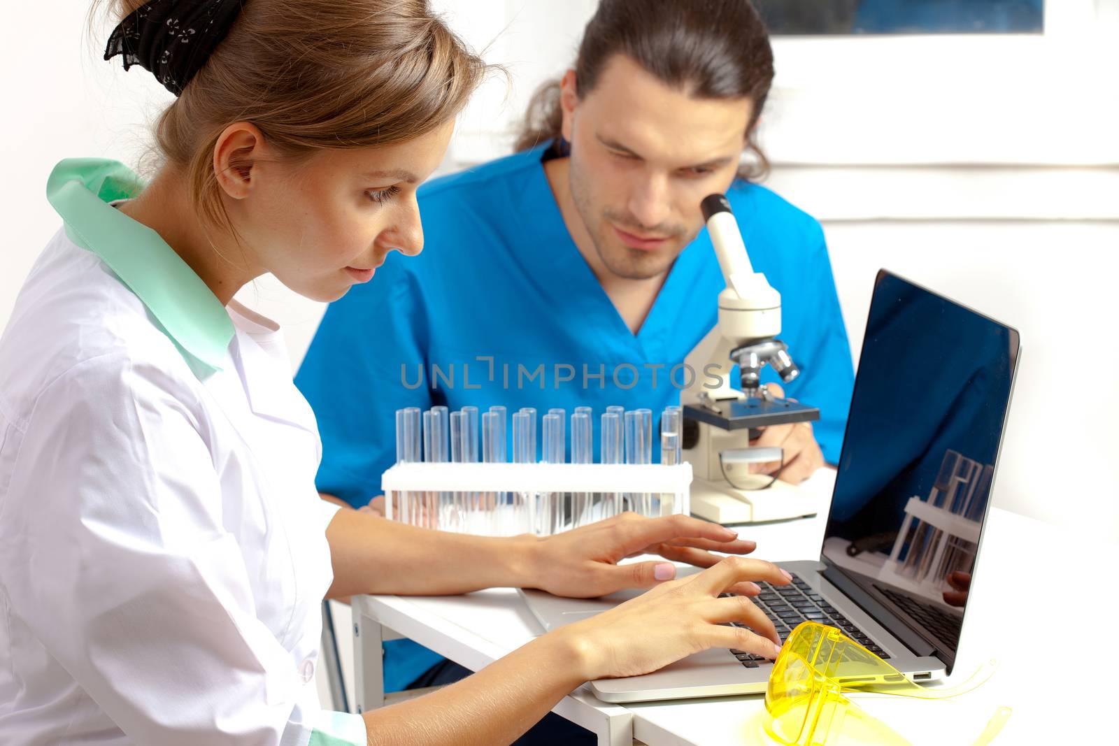 Doctor enters data into the computer, a researcher looking through a microscope in the background