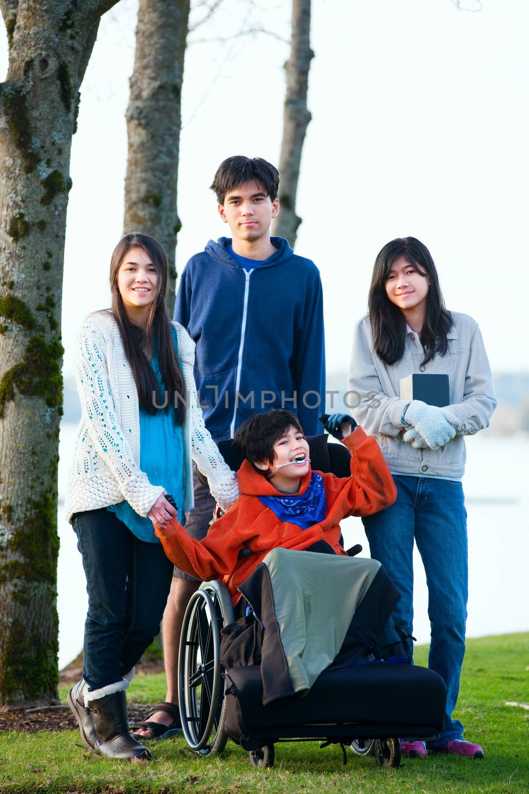 Disabled little boy in wheelchair surrounded by brother and sisters at lakeside. Child has cerebral palsy and children are all biracial