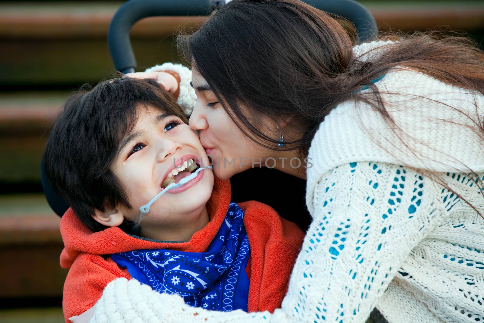 Big sister kissing disabled little brother seated in wheelchair  by jarenwicklund