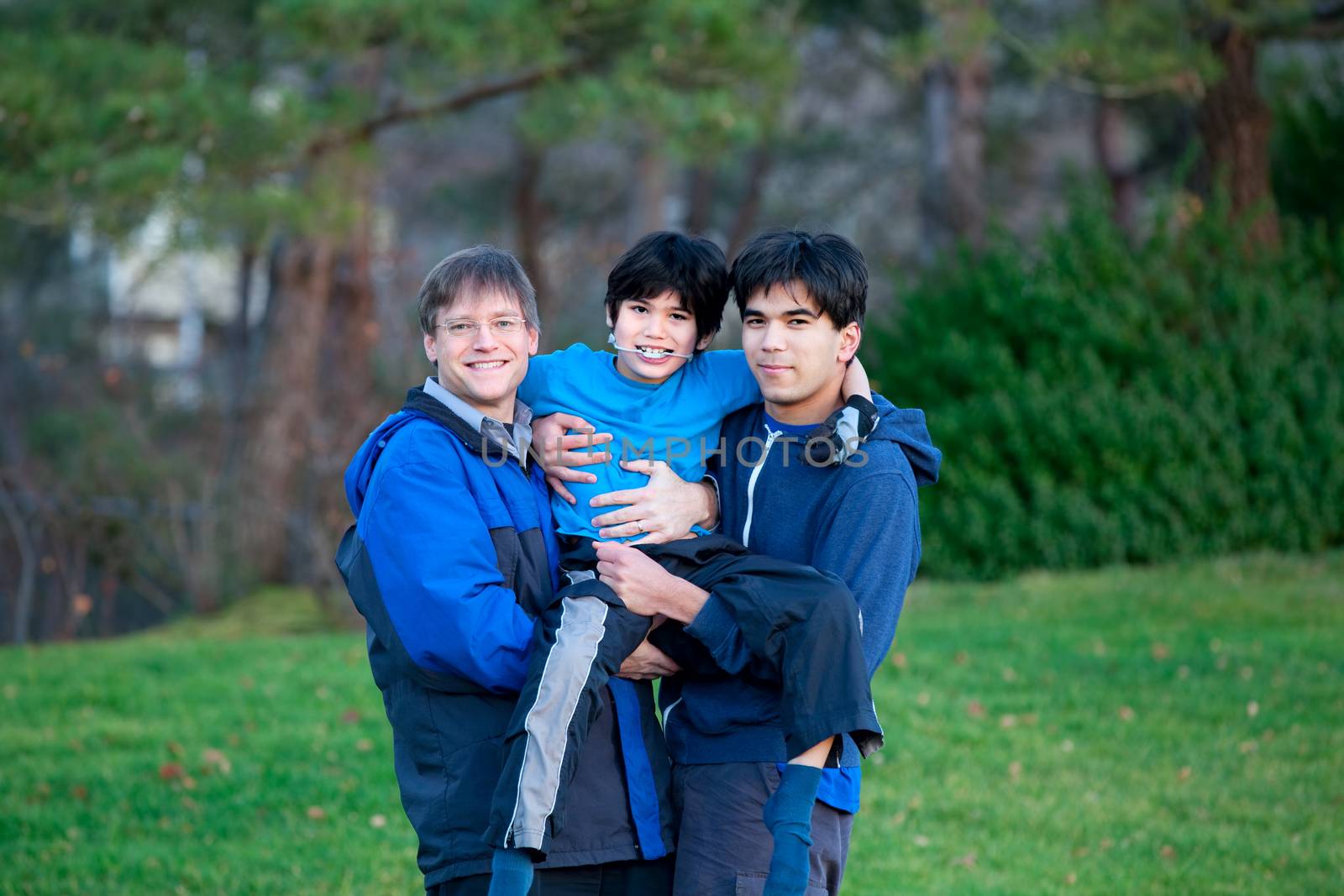 Disabled biracial child carried by his father and brother, at park.