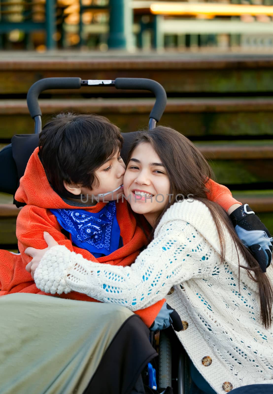 Disabled little boy kissing his big sister on cheek while seated in wheelchair