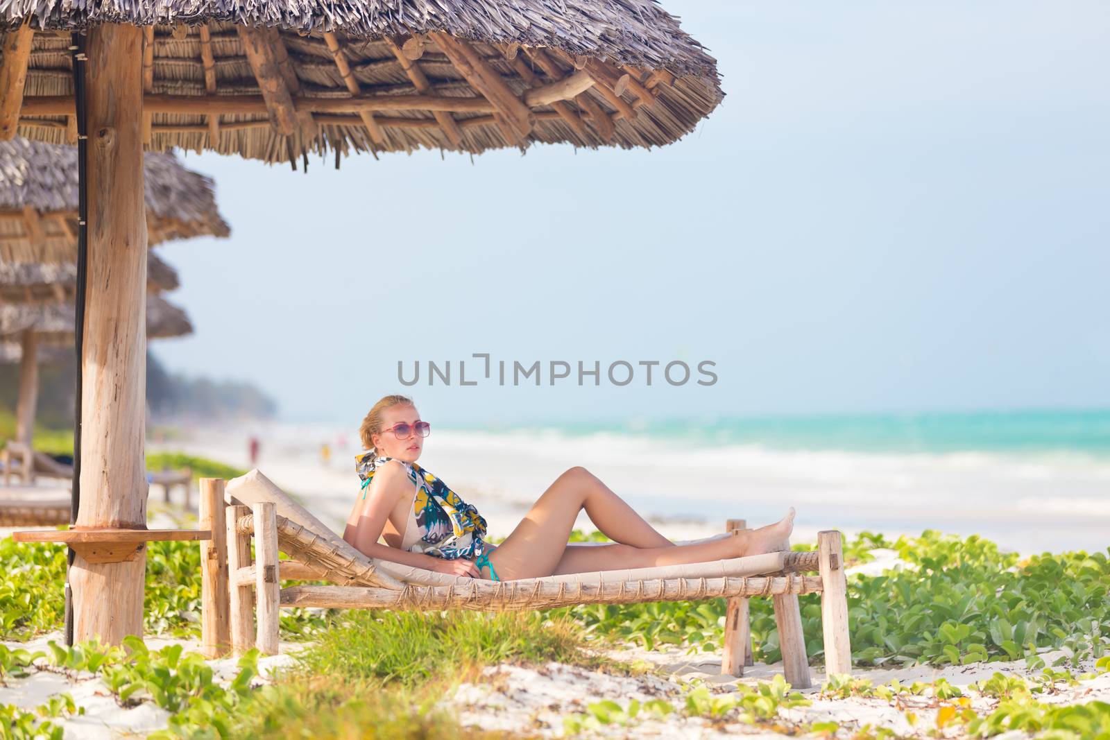 Women sunbathing on dack chair under wooden umbrella on stunning tropical beach. Kiteboarders turquoise blue lagoon of Paje beach, Zanzibar, Tanzania in the background.