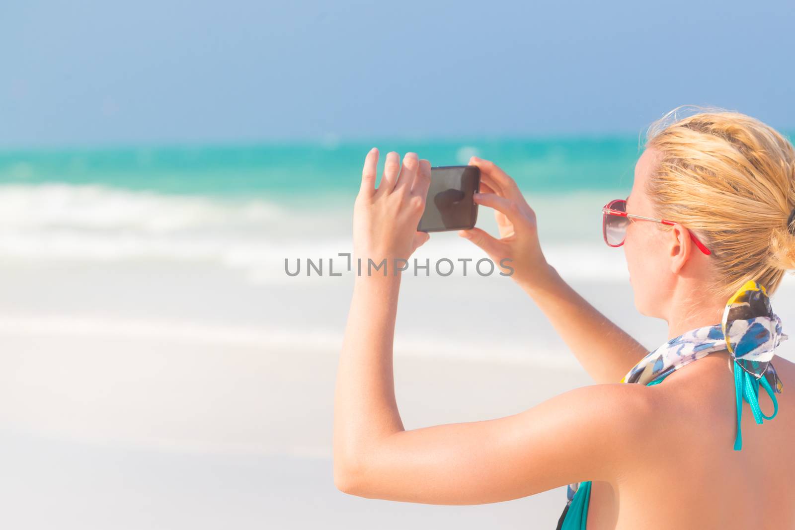 Blonde caucasian woman taking photo of blue tropical beach. Beautiful caucasian model  wearing turquoise swimsuit and colorful scarf on vacations on picture perfect Paje beach, Zanzibar, Tanzania.