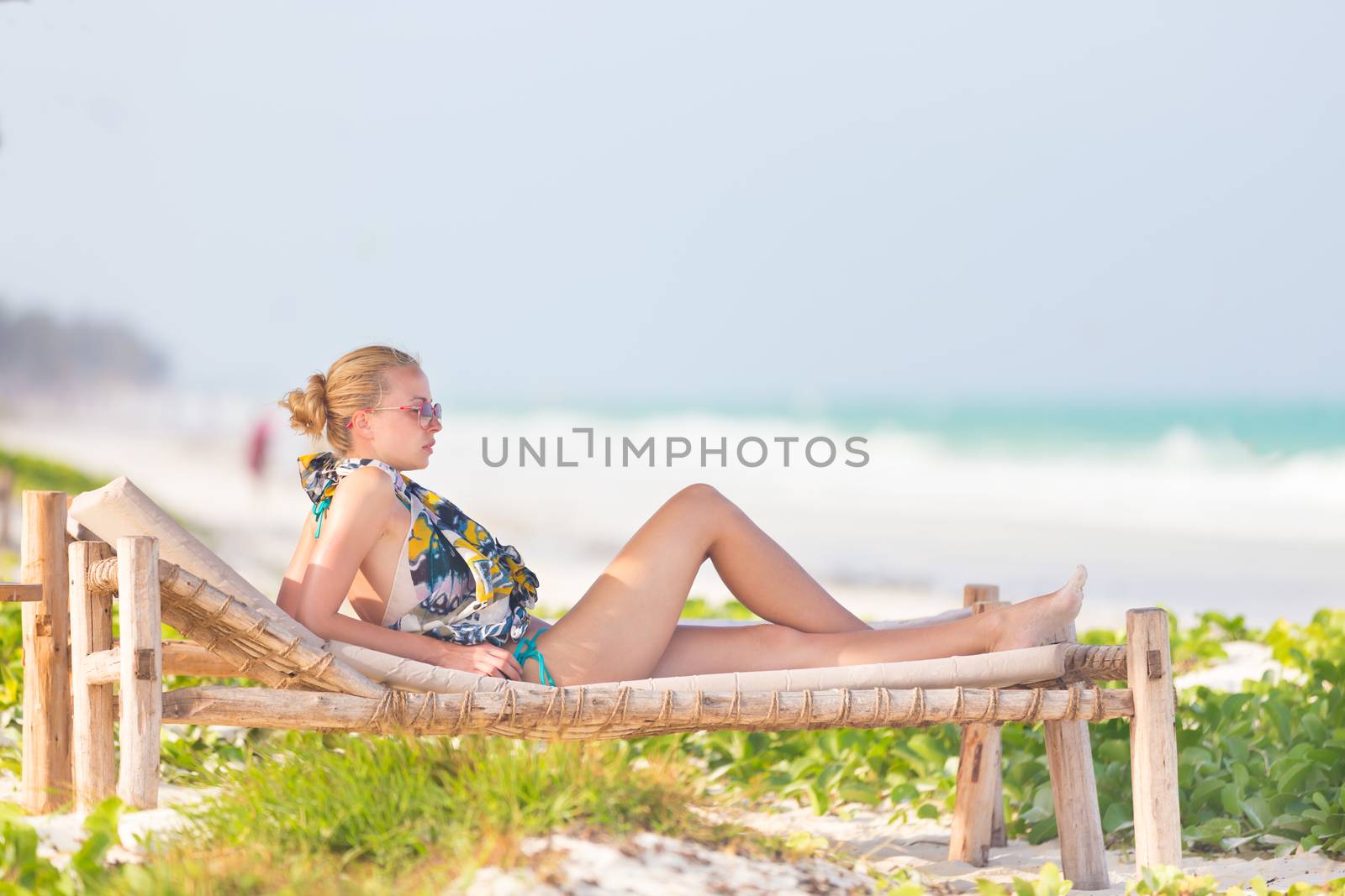 Women sunbathing on dack chair under wooden umbrella on stunning tropical beach. Kiteboarders turquoise blue lagoon of Paje beach, Zanzibar, Tanzania in the background.