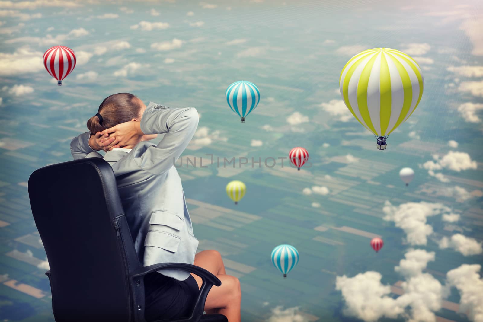 Businesswoman sitting with her hands clasped behind her head. Aerial view of vast crop acreage with a few air balloons above as backdrop