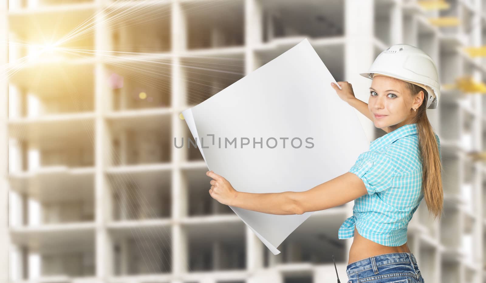 Woman in hard hat holding blank drawing sheet in front of building under construction, looking, at camera, smiling