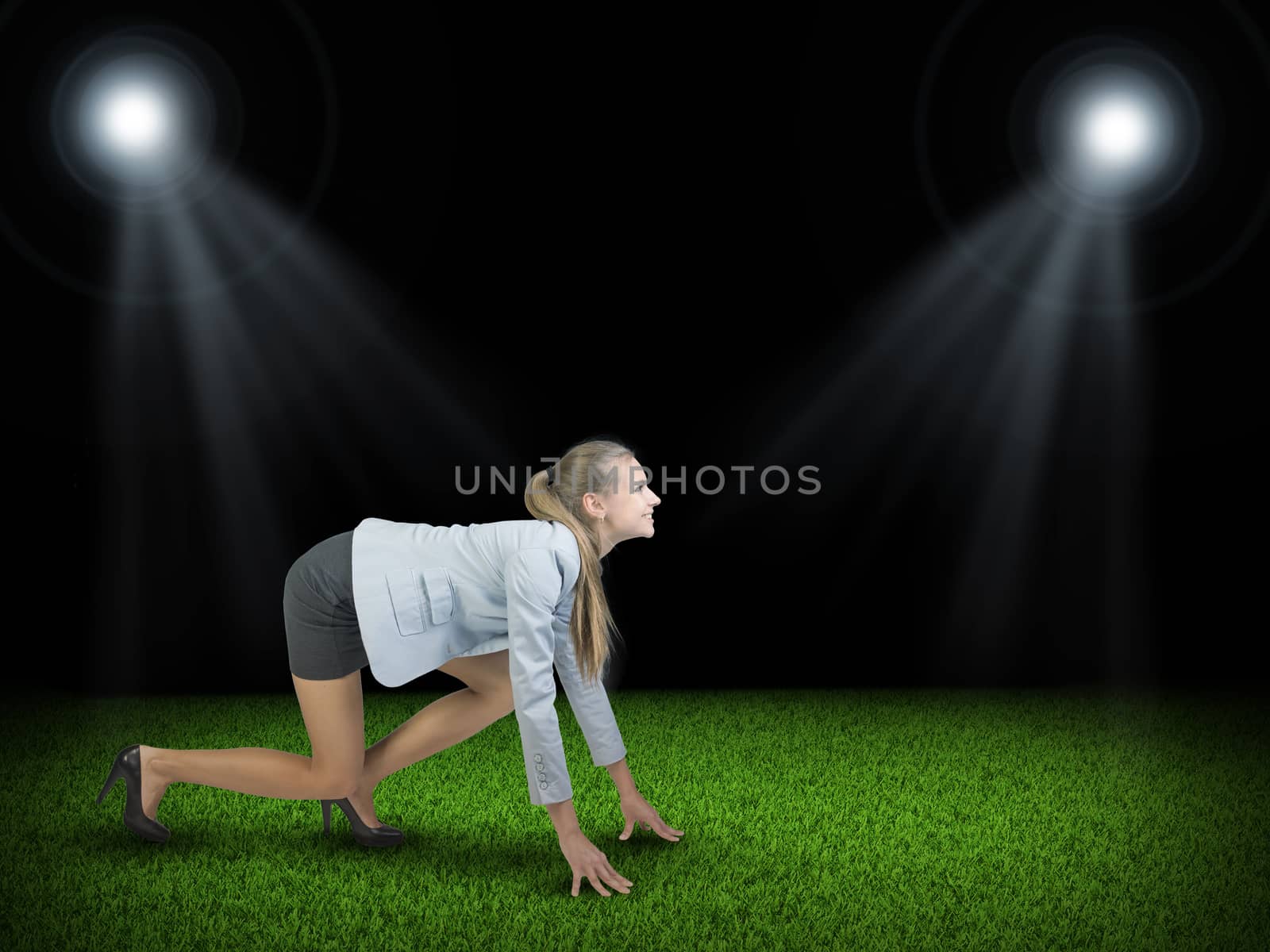 Businesswoman standing in running start pose at green grass court illuminated by two spotlights, looking ahead, smiling