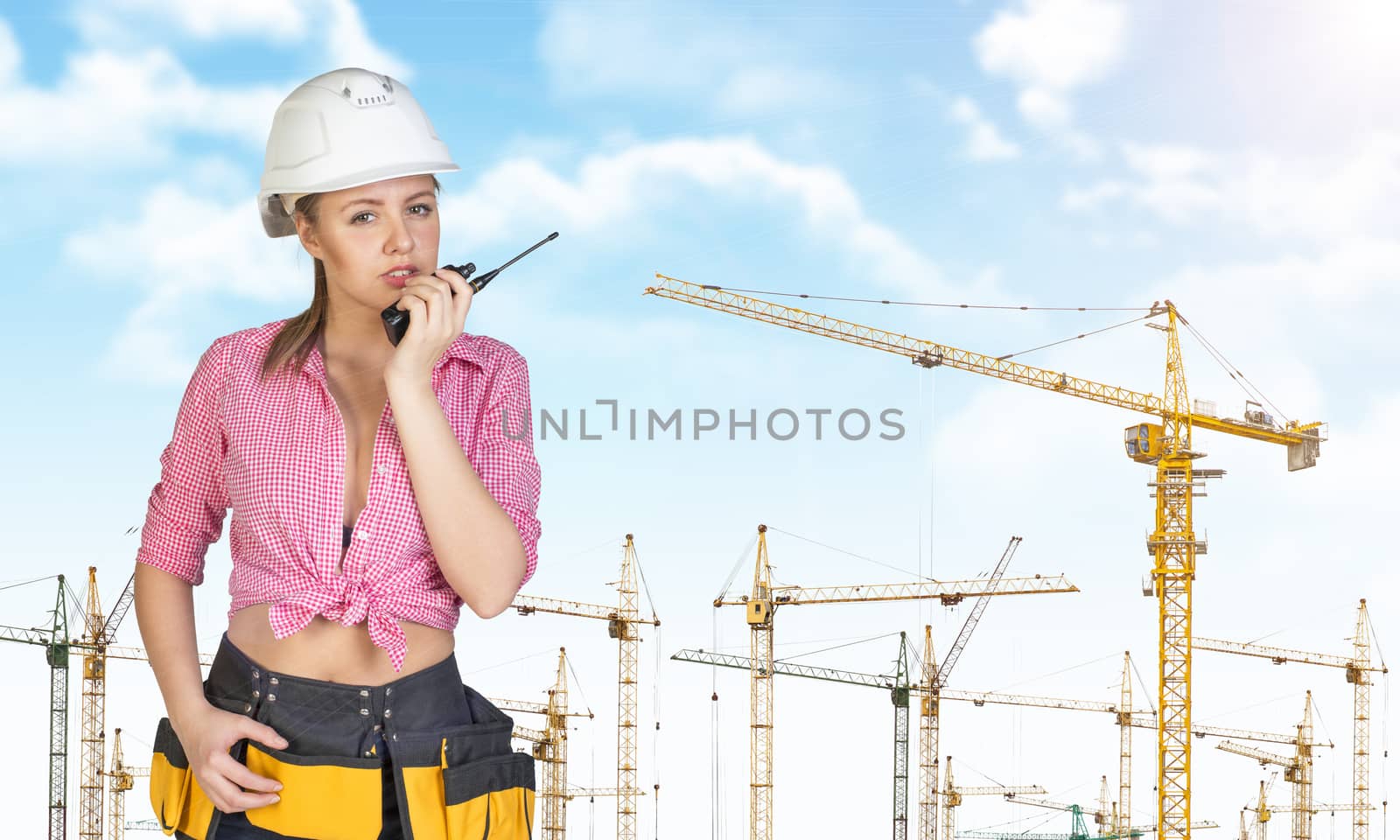 Woman in hard hat and tool belt talking on walkie talkie, looking at camera. Sky, clouds and tower cranes as backdrop