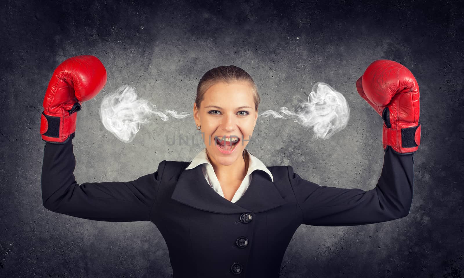 Businesswoman in boxing gloves posing with her arms up, looking at camera, shouting, smoke from her ears. Raw concrete wall as backdrop