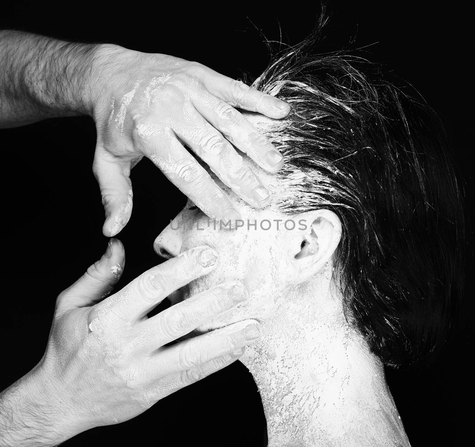 Black and white portrait of human hands working with woman with clay on face on black background