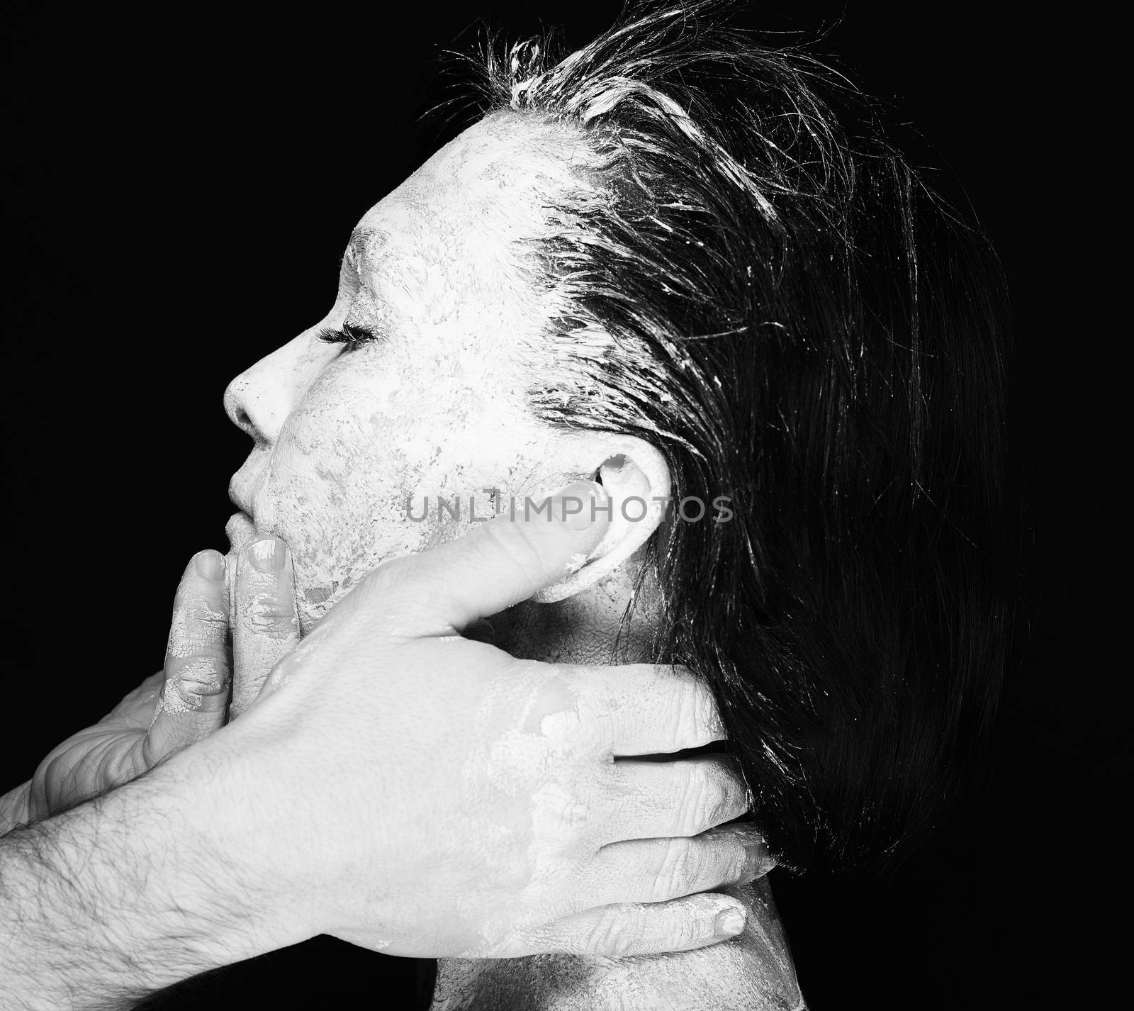 Black and white portrait of human hands working with woman with clay on face on black background