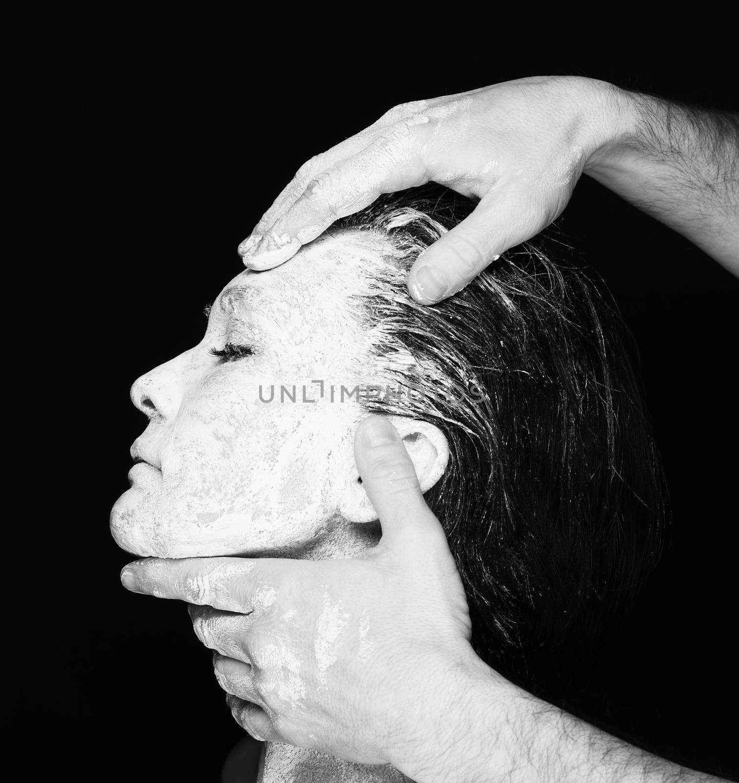 Black and white portrait of human hands working with woman with clay on face on black background