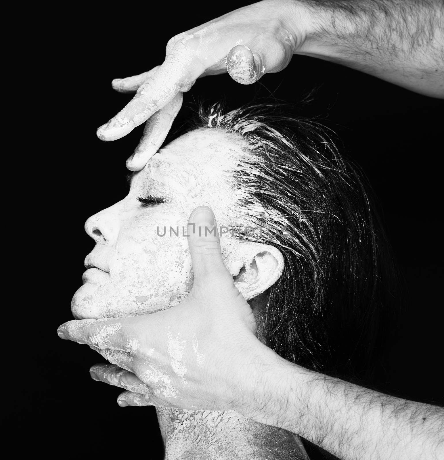 Black and white portrait of human hands working with woman with clay on face on black background