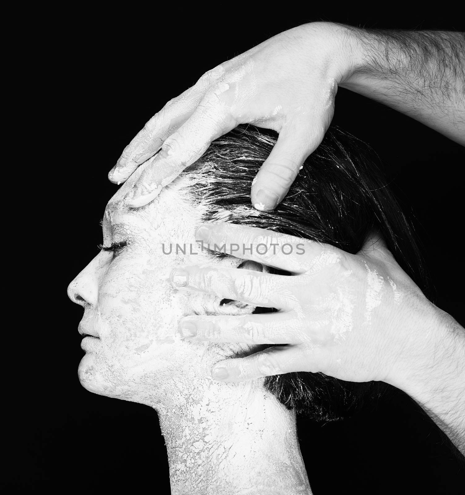 Black and white portrait of human hands working with woman with clay on face on black background