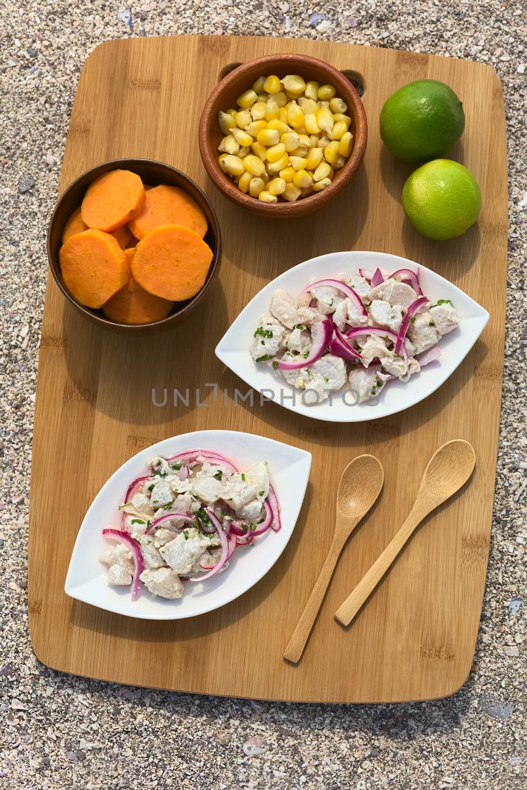 Overhead shot of Peruvian ceviche (raw fish and red onion marinated in lime juice with garlic, salt and coriander) served with sweet potato slices and cob kernels on wooden board on sand photographed with natural light