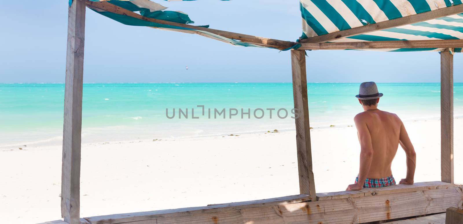Man hiding in shade from burning tropical sun on picture perfect tropical sandy beach. Summer leisure. Shot from back.
